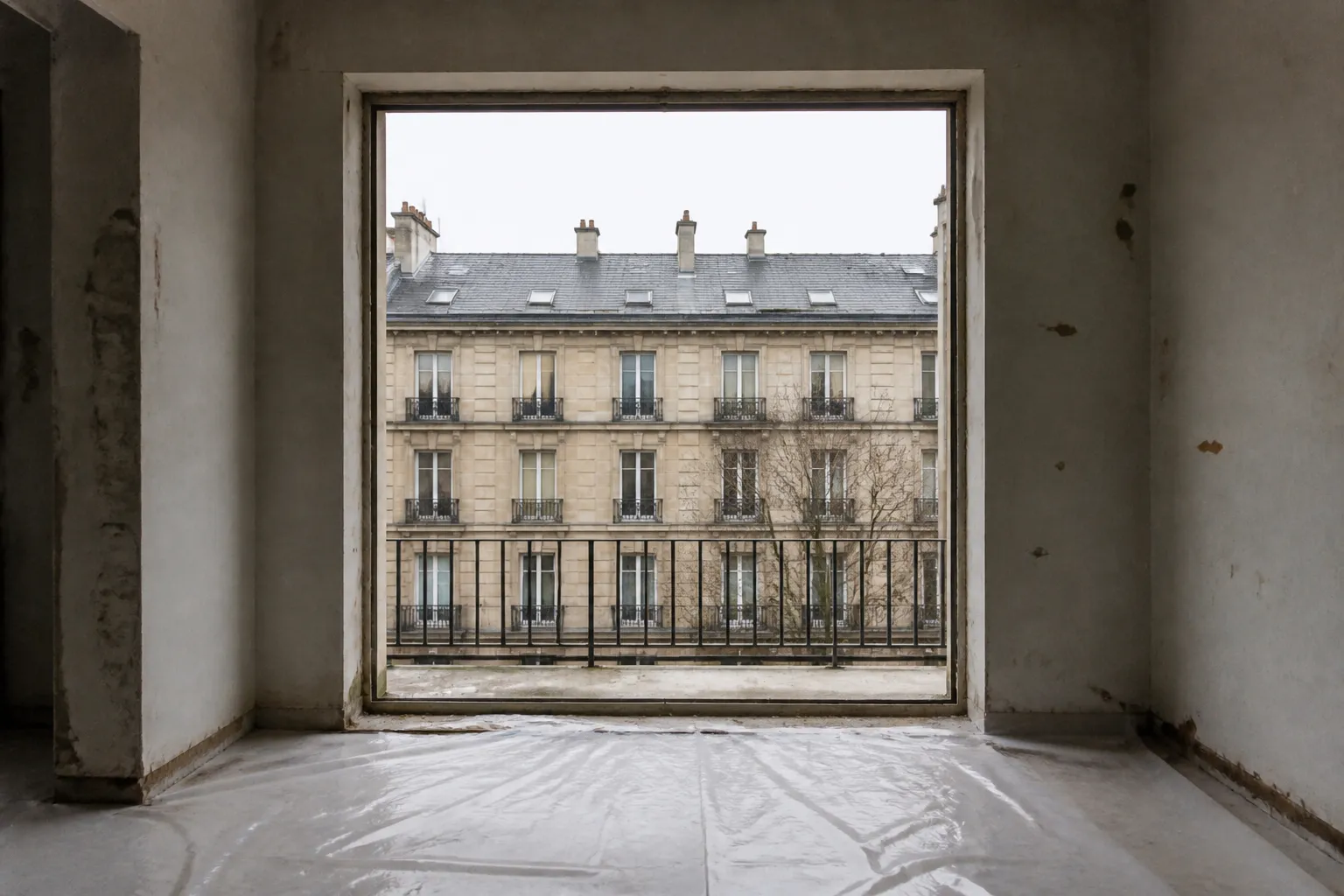 Baie vue depuis l’intérieur d’un logement en travaux, fenêtre déposée : l’ouverture est libre sur la rue et sur la façade d’en face, le tableau est brut, un film de protection couvre le sol