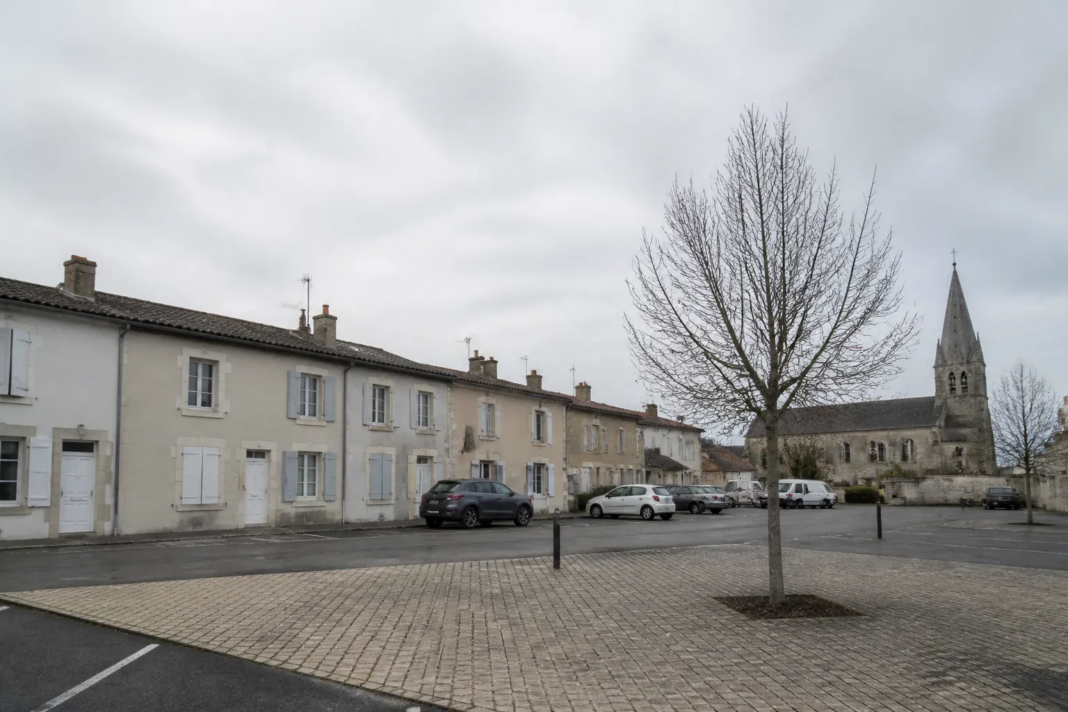 Place d’un petit bourg de plaine vue depuis son bord : maisons mitoyennes à un étage aux enduits clairs et volets simples, clocher de pierre en arrière-plan, ciel couvert