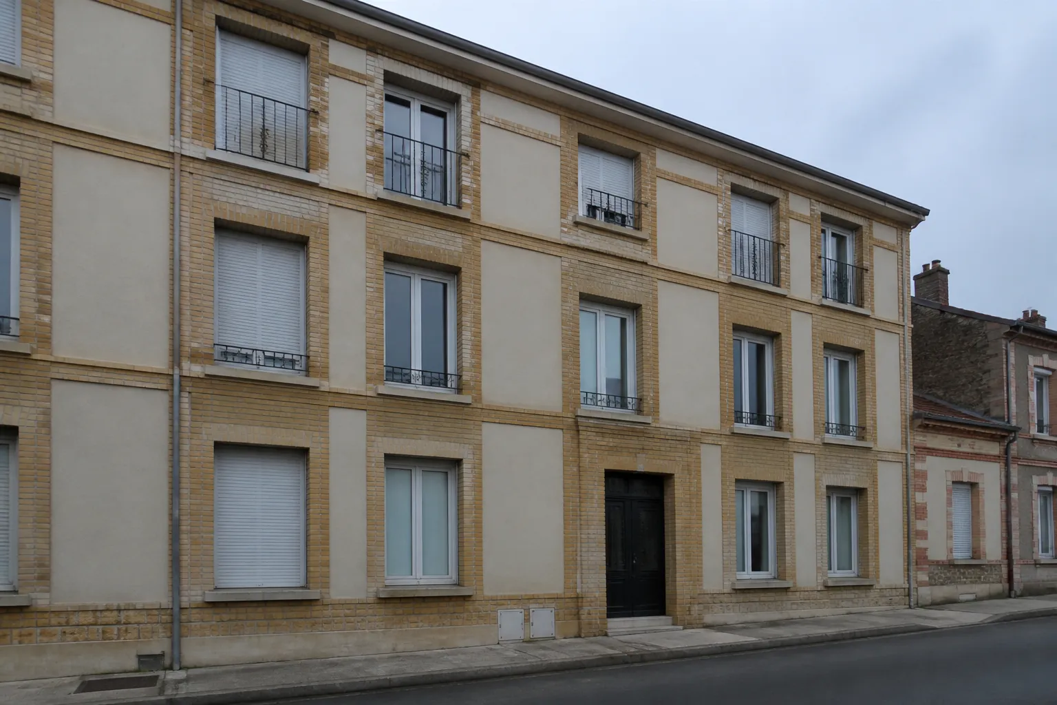 Façade d’immeuble des années 1920 vue depuis le trottoir d’en face : trame régulière de baies verticales, encadrements et bandeaux de brique de parement sur enduit clair, appuis saillants, ciel couvert