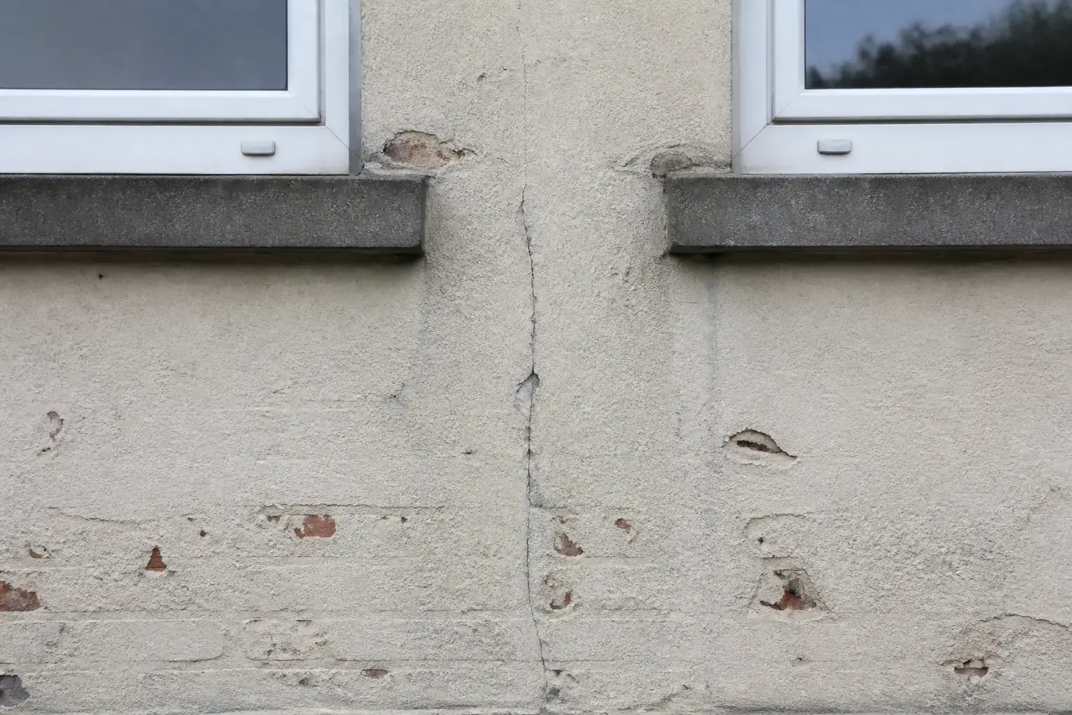 Fissure verticale fine courant dans l’allège d’une façade entre deux fenêtres, sur un enduit clair vieilli qui laisse deviner la brique, avec un léger décollement en bordure