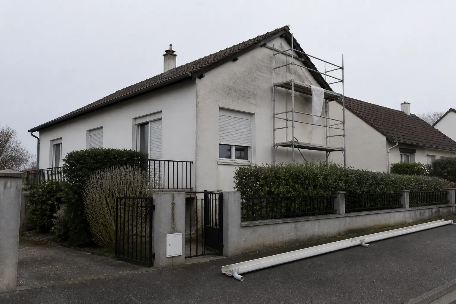 Maison individuelle de lotissement des années 1970-1980 vue depuis la rue, enduit clair et toiture de tuiles, échafaudage monté sur un pignon et gouttière déposée au sol
