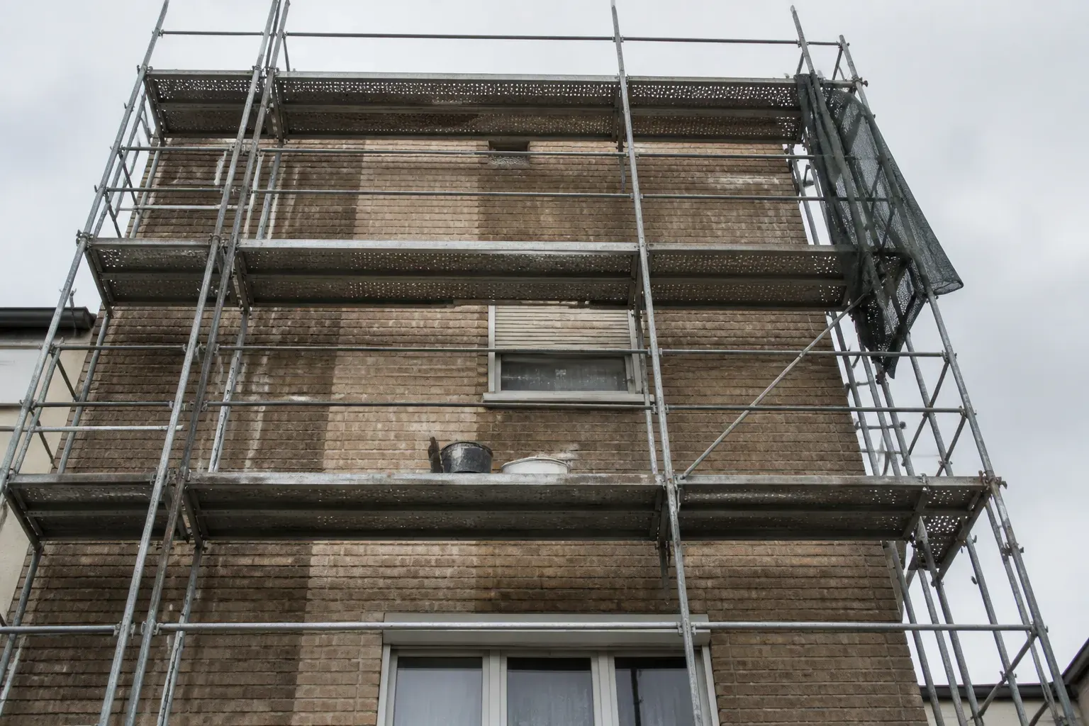 Façade de brique de parement en cours de ravalement sous échafaudage tubulaire : une bande verticale nettoyée et rejointoyée tranche nettement avec la maçonnerie encore encrassée